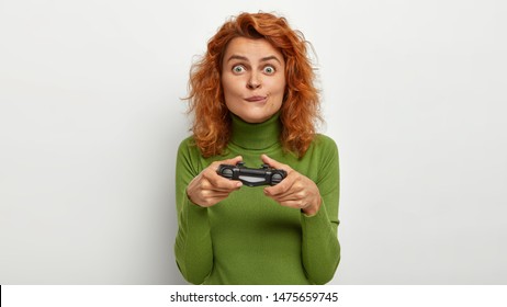 Gaming, hobby, excitement concept. Ginger female plays video game, holds joystick, purses lips and looks with interest, feels crazy, rests at home. Teenage girl with playstation or game controller