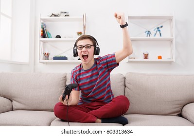 Gaming Video Games Concept - Excited Teenage Boy Playing Football Game With Joystick And Headphones, Enjoying Win While Sitting On Sofa In Living Room At Home