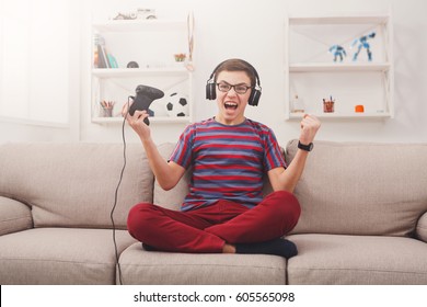 Gaming Video Games Concept - Excited Teenage Boy Playing Football Game With Joystick And Headphones, Enjoying Win While Sitting On Sofa In Living Room At Home