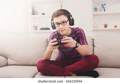 Gaming Video Games Concept - Teenage Boy Playing Football Game With Joystick And Headphones, Enjoying Sitting On Sofa In Living Room At Home