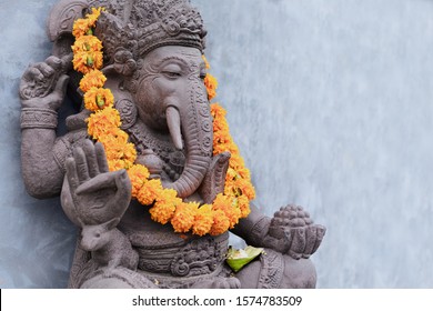 Ganesha Sitting In Meditating Yoga Pose In Front Of Hindu Temple. Decorated For Religious Festival By Orange Flowers Garland, Ceremonial Offering. Balinese Travel Background. Bali Island Art, Culture.