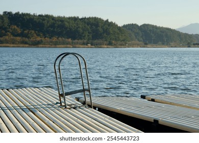 Gangneung, South Korea - November 3rd, 2024: A close-up of a metal ladder on a wooden dock at Hyang Lake, with calm waters and a forested shoreline in the background under a clear autumn sky. - Powered by Shutterstock