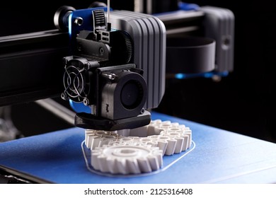 gantry with x-carriage and print head of a FDM-3D-printer that produces white helical gears on blue print bed. dark surrounding. selective focus. additive manufacturing concept