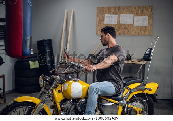 In the garage, a car\
mechanic sits on a yellow motorcycle that he has repaired. Pumped\
up stylish guy.