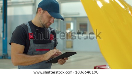 In a garage a mechanic using the tablet makes the oil and engine check to the car smiles. Concept of: security, safety, insurance and professionalism, revisions, technology.