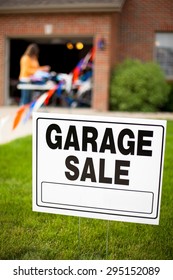 Garage Sale Sign On The Front Yard Of A Suburban Home With A Woman Looking At Items On A Table.