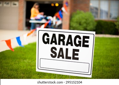 Garage Sale Sign On The Front Yard Of A Suburban House With A Woman Looking At Items On A Table.