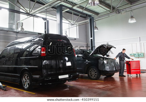 Garage work time, mechanic takes tools out of box.\
Black SUV in garage on foreground, mechanic near car with open hood\
and taking tools from box on background. SUV service maintenance in\
garage