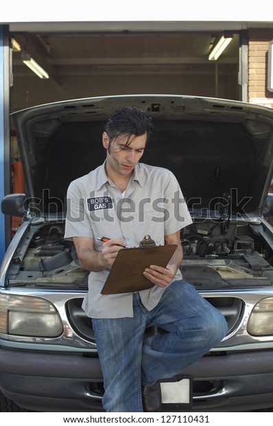 Garage worker writing on clipboard while leaning on\
an open car