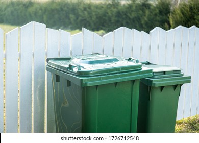 Garbage Bin With Waste Behind The White Wooden Fence Outside Of The Yard. Trash In Green Plastic Container.  Sorting The Separate Waste. Responsible Environmental Attitude. Reuse Of Different Trash.