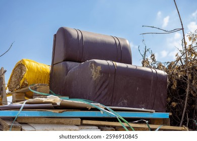 Garbage dump. An old brown leather chair with visible damage, abandoned on a pile of garbage and waste outdoors under a blue sky, symbolizing decay and waste.