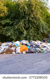 Garbage is stacked high in an open space surrounded by trees. The area shows signs of neglect with various items scattered among the waste.