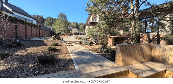 A garden area includes a stone path with small trees and shrubs. Houses are visible in the background. Sunlight shines on the scene. The setting appears welcoming and green.