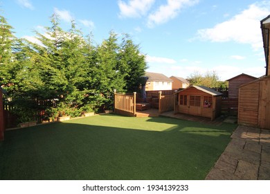 Garden With Astroturf Lawn With Wooden Deckig Area, Childrens Play House, Hut, Flowers And Trees.