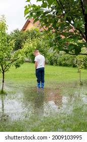 The Garden Is Flooded. Consequences Of Downpour, Flood. Rainy Summer Or Spring.The Child Stands In The Garden In The Water, Feet In Rubber Boots