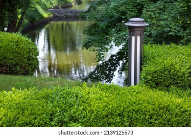 Garden Ground Lantern Of Street Lighting In Deciduous Boxwood Bushes Near A Pond With Trees On A Summer Day, Landscaping With Lighting Fixtures Objects.