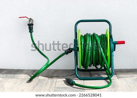 A garden hose connected to a faucet protruding from a building against a white facade.
