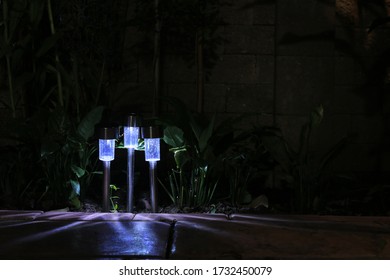 Garden LED Lamp Lighting. Small Solar Lighting Shining In A Garden And Casting Shadow In Multiple Direction. Cool Light Temperature For Modern Hi Tech Landscape Design. 