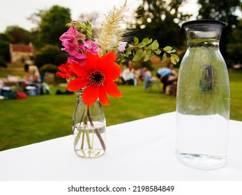 A Garden Party In An English Country Garden. In The Foreground Is A Water Bottle And A Flower Arrangement – Behind, Guests Enjoy Their Picnics On Folding Tables And Chairs.