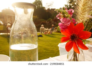 A Garden Party In An English Country Garden. In The Foreground Is A Water Bottle And A Flower Arrangement – Behind, Guests Enjoy Their Picnics On Folding Tables And Chairs.