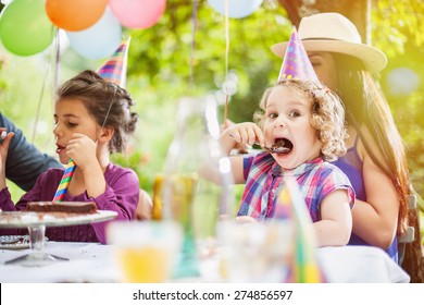 Garden Party Family Sharing The Birthday Cake. The Three Year Old Is Enjoying Her Piece, The Garden Is Decorated With Balloons And Colors Are Bright