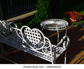 Garden White Wrought Iron Furniture In The Summer Garden. In The Foreground Is A Sofa With A Large Heart On The Back, Opposite It Is A Small Table. Close Up. Evening Lighting. 