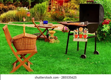 Garden Wooden Furniture, Picnic Hamper Basket, BBQ Grill, Sign Welcome, Wine Glasses On The Table, Plants, Trees and House In The Background. Backyard  BBQ Grill Party Or Picnic Concept