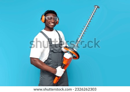 garden worker in uniform holding electric brush cutter on blue isolated background, african american man in overalls goggles and construction headphones with gardening tool Stockfoto © 