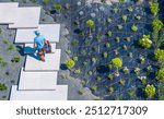 A gardener carrying tools walks along stepping stones in a contemporary landscaped garden filled with various plants and drip irrigation system.