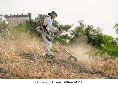 A gardener, dressed in full white protective gear and a face shield, is seen cutting dry grass on a slope with a lawnmower. The dust raised by the trimmer highlights the intensity of the work. - Powered by Shutterstock