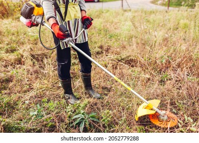 Gardener mowing weeds with brush cutter in fall meadow. Worker trimming dry grass with manual gasoline trimmer with metal blade disk. Farmer wearing boots