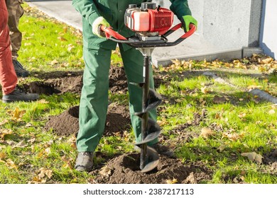 Gardener using tools hand-held soil hole drilling machine or portable manual earth auger for prepare the soil for planting shrubs or trees.