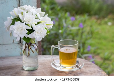 Gardenia Bouquet In A Glass Bottle And A Cup Of Tea Put On The Wooden Table Against Green Fresh Plant In The Home Garden.