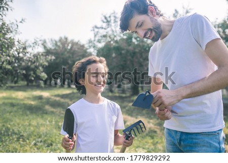 Gardening. Dark-haired boy and his dad holding gardening equipment and smiling