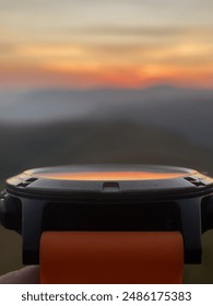 A Garmin Forerunner watch with an orange silicone band faces a colorful sunset. The watch has a black bezel and a round display.