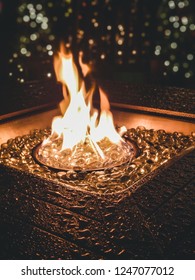 Gas Flames Dance In An Outdoor Fire Pit Lined With Glass Pebbles. Decorated Christmas Trees In The Background. Rocking Chairs Around A Fireplace Outside Wet From Rain.