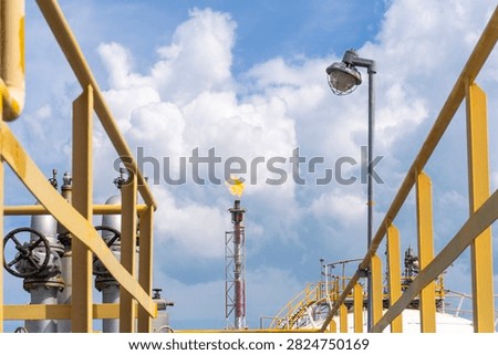 Gas flaring from a flare stack visible between yellow railings at a petrochemical plant, with bright flames burning against a blue sky with white clouds.