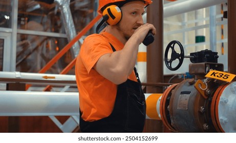 Gas plant worker operating pipeline valve using walkie talkie. Industrial worker wearing hard hat and hearing protection is operating pipeline valve and speaking into walkie talkie