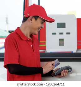 Gas Station Worker In Red Uniform Stand Smiling, Swipe Mockup Credit Card Via Payment Terminal. Cash, Price And Volume Readout Display On Petrol Pump Display Screen.