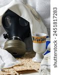 Gasmask, partially concealed by white prayer talit, next to a Kaddish cup on a table with matzah bread. Wine holiday matzoth celebration matzoh jewish passover bread. Traditional elements and survival