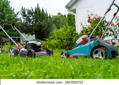 Gasoline And Battery Electric Lawn Mowers In The Garden Against The Backdrop Of A Blooming Garden, Old And New Grass Mowing Technologies