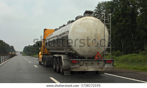 Gasoline and oil transportation, old chrome gray\
barrel fuel tank semi trailer with American truck on suburban\
highway road at summer day on green forest on roadside background,\
rear side view closeup