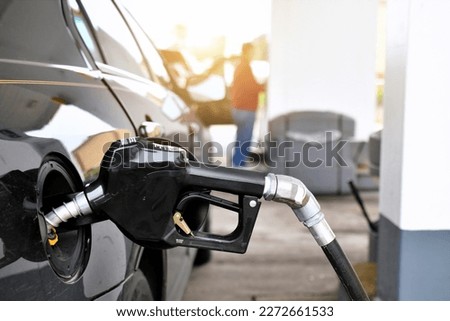 A gasoline pump handle in a gas tank of vehicle with another customer at gas pump in background