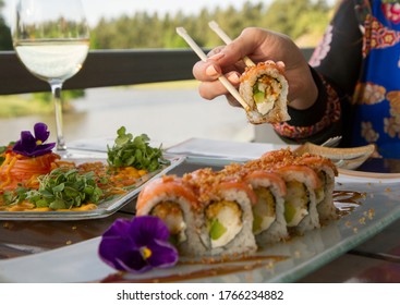 Gastronomy. Elegant Sushi Restaurant. Woman Dining Outside With A Natural Lake Background. Young Female Hands Holding A Raw Salmon Sushi Roll With The Chopsticks.