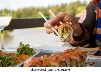 Gastronomy. Elegant Sushi Restaurant. Young Female Hands Holding A Raw Salmon Sushi Roll With The Chopsticks. Woman Dining Outside With A Natural Lake Background. 