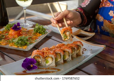 Gastronomy. Elegant Sushi Restaurant. Young Female Hands Holding A Raw Salmon Sushi Roll With The Chopsticks. Woman Dining Outside With A Natural Lake Background.