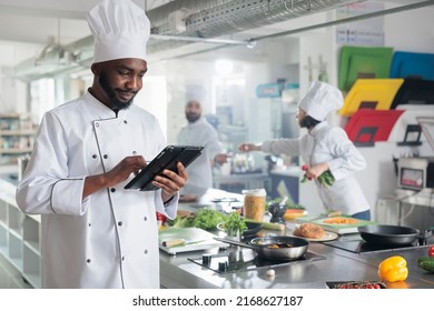 Gastronomy expert with handheld device standing in restaurant professional kitchen while looking for dinner recipes. Head cook with tablet brainstorming garnish ideas for gourmet cuisine meal. - Powered by Shutterstock