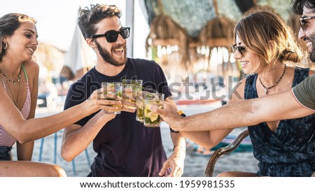Gathering of millennial friends clinking with cocktail glasses at the beach cafe having fun together in the summer. Concept of young people drinking alcohol together and interacting with each other