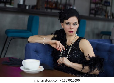 Gatsby style young beautiful girl in the image of the 1920s in a black dress in long black gloves in a black boa on her shoulder sits at a table