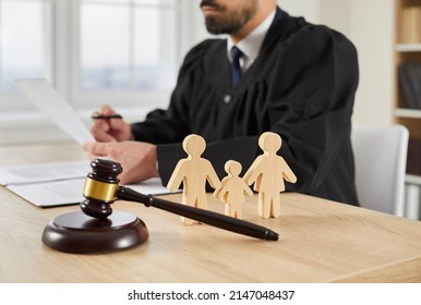 Gavel And Little Wooden Figures Of Husband, Wife And Child Up Close On Table In Courthouse, And Judge Reading Divorce Settlement In Background. Family Law, Court Trial, Parent Getting Custody Concept